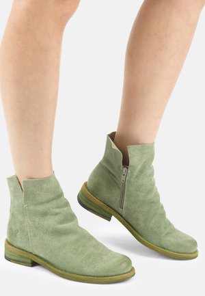 Beine, die hellgrüne Wildleder-Stiefeletten mit seitlichem Reißverschluss und niedriger Holzsohle tragen, vor weißem Hintergrund gezeigt.