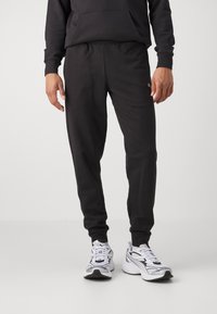 Sorte sweatpants med elastisk talje og manchetter, lavet af et glat stof. Sammen med grå og hvide træningssko.