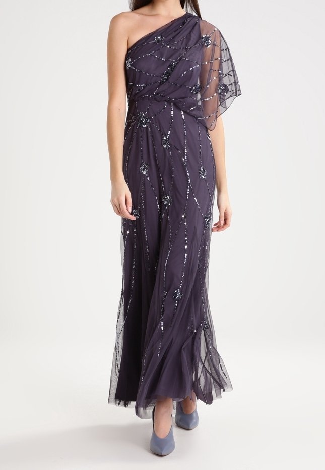 Femme portant une robe de soirée asymétrique violette foncée avec des motifs floraux en sequins et un tissu transparent, associée à des talons violets clairs.