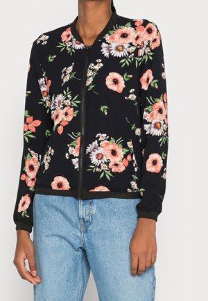 Femme portant une veste noire zippée avec des motifs floraux roses et blancs et un jean bleu clair sur un fond uni.