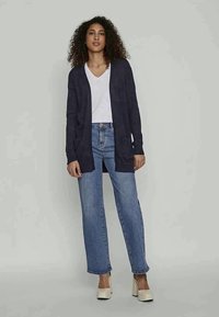 Cardigan en tricot bleu marine avec une coupe ouverte, équipé de manches longues et de poches latérales, associé à un t-shirt blanc à col en V et un jean taille haute.