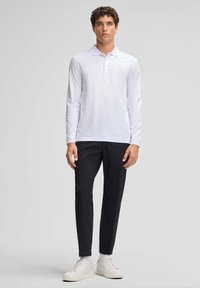 Polo a maniche lunghe bianco con una consistenza morbida, abbinato a pantaloni neri slim-fit e sneakers bianche, in piedi contro uno sfondo neutro.