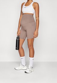 Frau in beigen Umstands-Radshorts und weißem Sport-BH, die eine schwarze Nike-Wasserflasche hält, trägt weiße Nike-Sneaker und Socken.