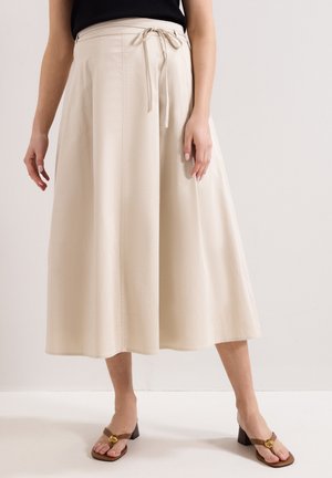 Beige wijde cropped broek met zijstrik, gedragen met een zwart topje en bruine sandalen met hak en gouden schelpdetaillering.