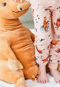 Barn i rosa byxor med häst- och fågeltryck står bredvid en stor brun plyschhäst-leksak på en vit yta.