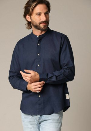 Chemise bleue marine à boutons avec col montant, manches longues et coupe décontractée. Fabriquée dans un tissu texturé, elle présente un détail de label sur le côté.