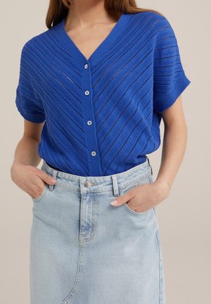 Kobieta w niebieskim, krótko rękawowym, zapinanym na guziki dzianinowym topie, włożonym w jasnoniebieską spódnicę dżinsową z wysokim stanem, z rękami w kieszeniach.