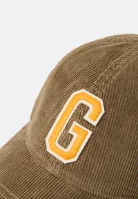 Gorra de pana marrón con una letra "G" bordada en color naranja en la parte frontal, con una superficie texturizada y detalles de costura.