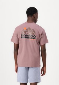 Helles mauve Baumwoll-T-Shirt mit einem grafischen Design von Bergen und Wellen in dunklen Farbtönen sowie einer orangen Sonne, kurzärmlig und mit Rundhalsausschnitt.