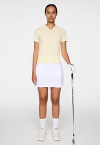 Vaaleankeltainen poolopaita valkoisella kauluksella, yhdistetty valkoiseen hameeseen. Valkoiset sukat ja kengät. Pitelee golfmailaa. Yksinkertainen muotoilu.