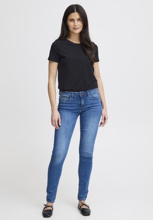 Černé tričko s krátkým rukávem spojené se středně modrými úzkými džíny. Modelka má na sobě černé baleríny; pozadí je jednobílé.