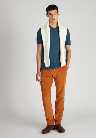 Męski zestaw składa się z turkusowego t-shirtu, kremowego swetra dzianinowego zarzuconego na ramiona oraz spodni z kordurowej tkaniny w kolorze rdzy. Całość dopełniają brązowe buty.