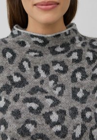 Vrouw in een grijze fuzzy trui met een zwart-wit luipaardprint en een hoge, losse halslijn.