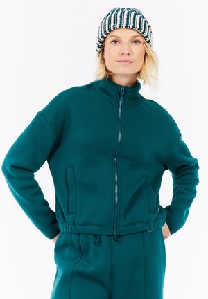 Vrouw in een turquoise zip-up jack en bijpassende broek met haar handen op de heupen, en een gestreepte gebreide muts.