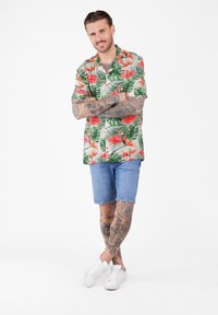 Mann mit Tattoos, der ein kurzärmliges Hemd mit Blumen- und Flamingo-Muster, Jeansshorts und weiße Sneakers trägt, steht mit verschränkten Armen.