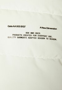 Vitt tyg med en quiltad textur som visar svart tryckt text, inklusive produktinformation och ett varumärkesuttalande, centrerat på materialet.