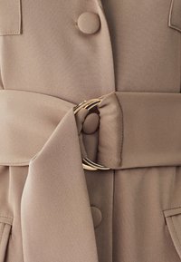 Beige Stoffkleid mit großen Knöpfen, einem Gürtel mit einer runden goldenen Schnalle und einer glatten Textur. Zwei vordere Taschen sind sichtbar.