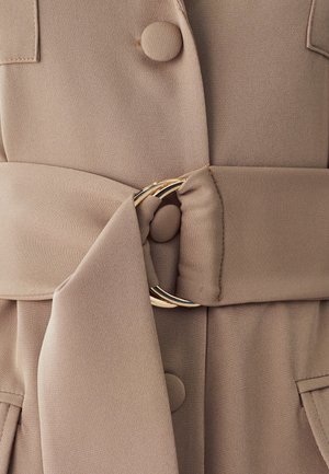 Beige stoffen kledingstuk met grote knopen, een riem met een cirkelvormige gouden gesp en een gladde textuur. Twee voorzakken zijn zichtbaar.