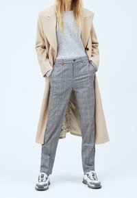 Beżowy długi płaszcz na szarym swetrze, w połączeniu z jasnoszarymi spodniami w kratę i białymi sneakersami. Wyróżnia się dopasowanym krojem i gładkimi fakturami.