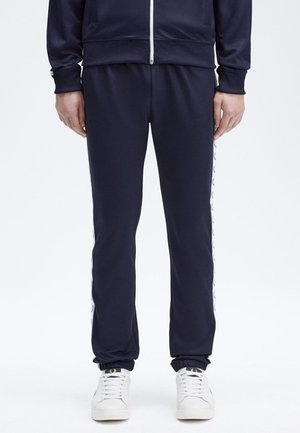 Homme portant un pantalon de survêtement bleu marine avec des bandes blanches sur les côtés et des baskets blanches, debout devant un fond uni.