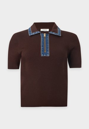 Femme debout portant un haut en maille marron à manches courtes avec un col bleu et un pantalon large foncé avec des chaussures noires.