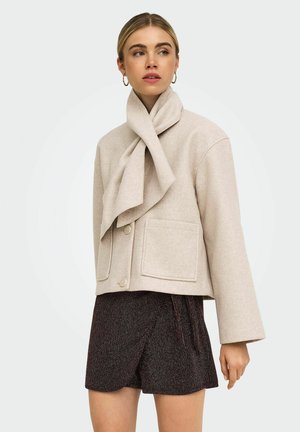 Femme porte une veste en laine beige clair avec de grandes poches avant et un col type écharpe, associée à un short texturé foncé.