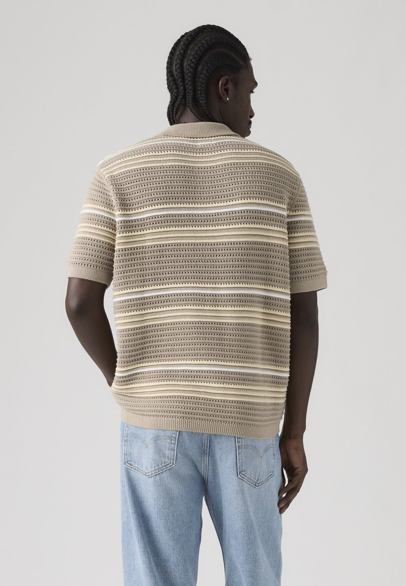 Personne aux cheveux tressés portant un pull en maille à manches courtes beige et blanc rayé et un jean bleu clair, vue de dos sur un fond uni.