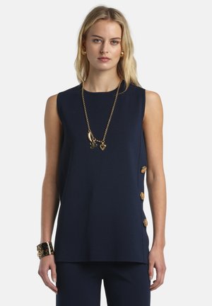 Mujer con top sin mangas azul marino con botones dorados en el lateral, collar con dije dorado, pendientes dorados y pulsera de carey.