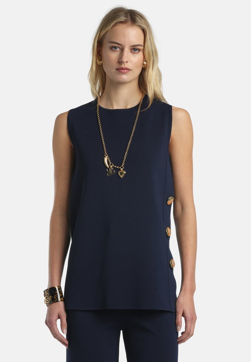 Femme portant un haut sans manches bleu marine avec des boutons dorés sur le côté, un collier à pendentif doré, des boucles d'oreilles dorées et un bracelet manchette en écaille.