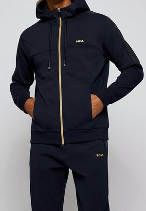 Homme portant un sweat à capuche zippé bleu marine et un pantalon assorti avec des logos "BOSS" dorés ainsi que des accents dorés sur la fermeture éclair et les cordons.