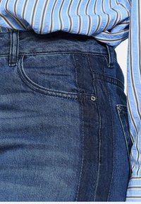 Donkerblauwe denim jeans met zichtbare voorzak en ton sur ton stiksel; gedragen met een lichtblauw gestreept overhemd met knopen.