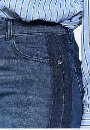 Jeans in denim blu scuro con tasche anteriori visibili e cuciture tono su tono; indossati con una camicia a righe blu chiaro con bottoni.