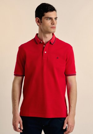 Camisa polo roja con cuello a rayas en navy, tapeta de dos botones y un pequeño logo en el lado izquierdo del pecho. Mangas cortas, tejido suave.