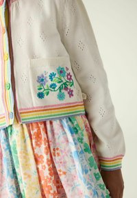 Cardigan bianco con una tasca caratterizzata da un ricamo floreale colorato, abbinato a un vestito con fantasia floreale multicolore. Polsini a righe arcobaleno.