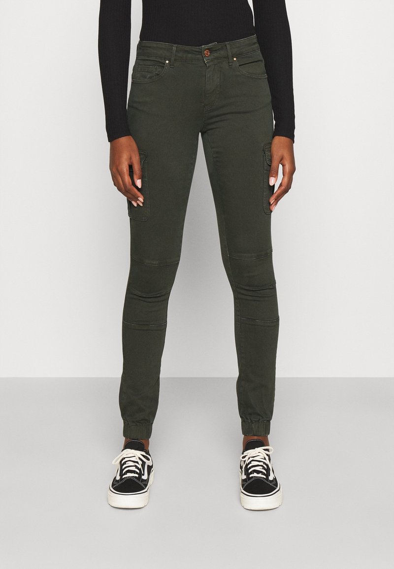 Personne portant un pantalon cargo vert foncé ajusté avec des coutures aux genoux, des poignets élastiques, des baskets noires et blanches, et un haut noir à manches longues.
