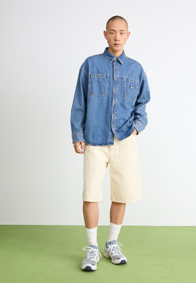 Camisa de mezclilla con botones y dos bolsillos en el pecho, pantalones cortos beige hasta la rodilla, calcetines blancos y zapatillas deportivas grises con acentos azules sobre un suelo verde.