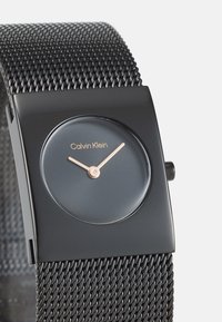 Orologio con bracciale in rete nera, cassa rettangolare, quadrante grigio, lancette in oro rosa e logo Calvin Klein. Design minimalista, texture elegante.