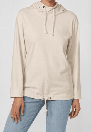 Top beige de manga larga con capucha y cordones en el cuello y el dobladillo, combinado con vaqueros azul claro, vista frontal desde la mitad del muslo hacia arriba.