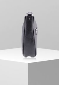 Czarna, teksturowana skórzana torba na ramię stojąca w pionie na białym cokole z widocznym srebrnym zamkiem błyskawicznym i metalowym pierścieniem.