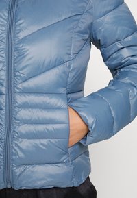 Blå puffjacka med horisontella och diagonala kviltade sektioner, med en hel frontdragkedja och handfickor på sidorna.