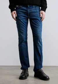 Vaqueros de mezclilla azul con corte recto, que presentan un diseño clásico de cinco bolsillos y un sutil desgaste. Combinados con mocasines negros.