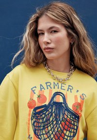Geel sweatshirt met de tekst "FARMER'S MARKET" en een illustratie van een blauwe mesh draagtas met rode tomatenaccenten, gecombineerd met een zilveren kettinkje.