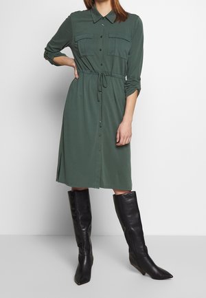 Femme portant une robe chemise à manches longues vert olive avec des boutons sur le devant, une taille à cordon et des bottes noires pointues montantes jusqu'aux genoux.
