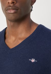 Pull en V bleu marine en tissu doux, présentant un tricot texturé et un petit logo brodé sur la poitrine gauche.