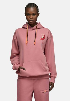Femme portant un sweat à capuche rose et un pantalon assorti avec un motif d'oiseau rouge sur la poitrine et des cordons orange à la capuche.