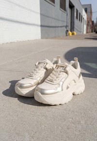 Ζευγάρι λευκά chunky sneakers με παχιές σόλες τοποθετημένα σε τσιμεντένιο πεζοδρόμιο δίπλα σε κτίριο με λευκούς και γκρι τοίχους.