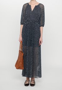 Robe maxi bleu marine avec imprimé floral blanc, manches transparentes et décolleté en V. Associée à un sac fourre-tout en osier marron et des chaussures plates noires.