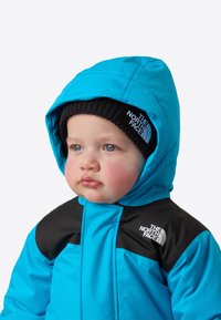Blå ytterjakke med hette, svarte skuldre og hvit logo. Barnet har på seg en svart strikkelue som fremhever ansiktstrekkene.