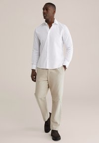 Wit langärmlig shirt met een slanke pasvorm, gecombineerd met beige broek en donkerbruine schoenen. De outfit heeft een strak, modern ontwerp.
