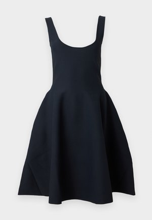 Femme aux cheveux foncés mi-longs, portant une robe noire sans manches, tenant un petit sac à main noir avec une bandoulière chaîne dorée.